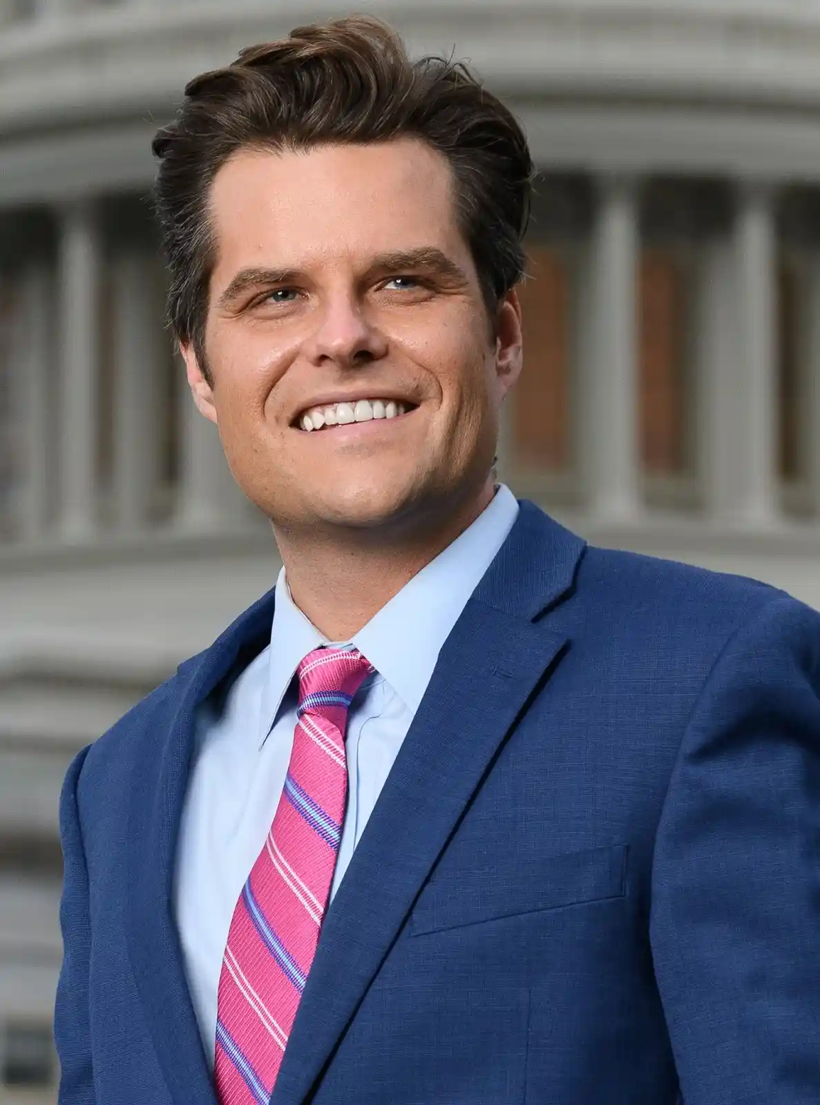 A smiling man in a blue suit and light blue shirt wears a bright pink tie with blue and white stripes, standing in front of…