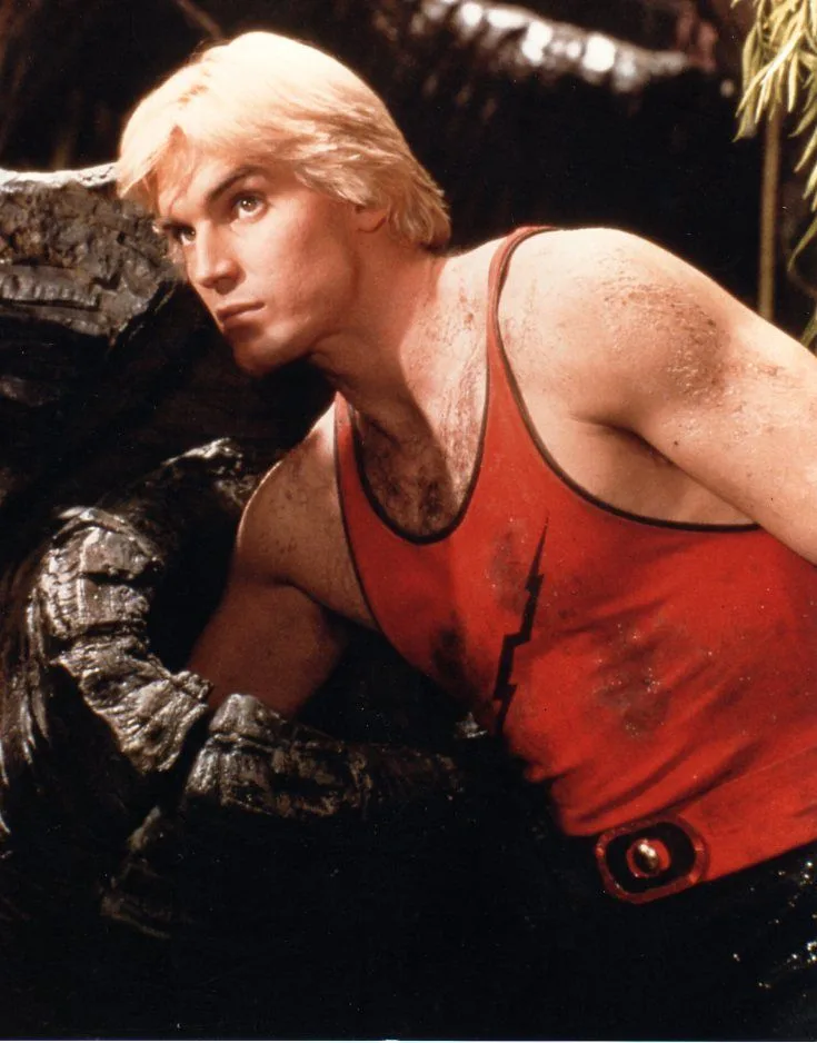 A man with blond hair and a red tank top with a black lightning bolt emblem is shown leaning against a tree trunk, covered…