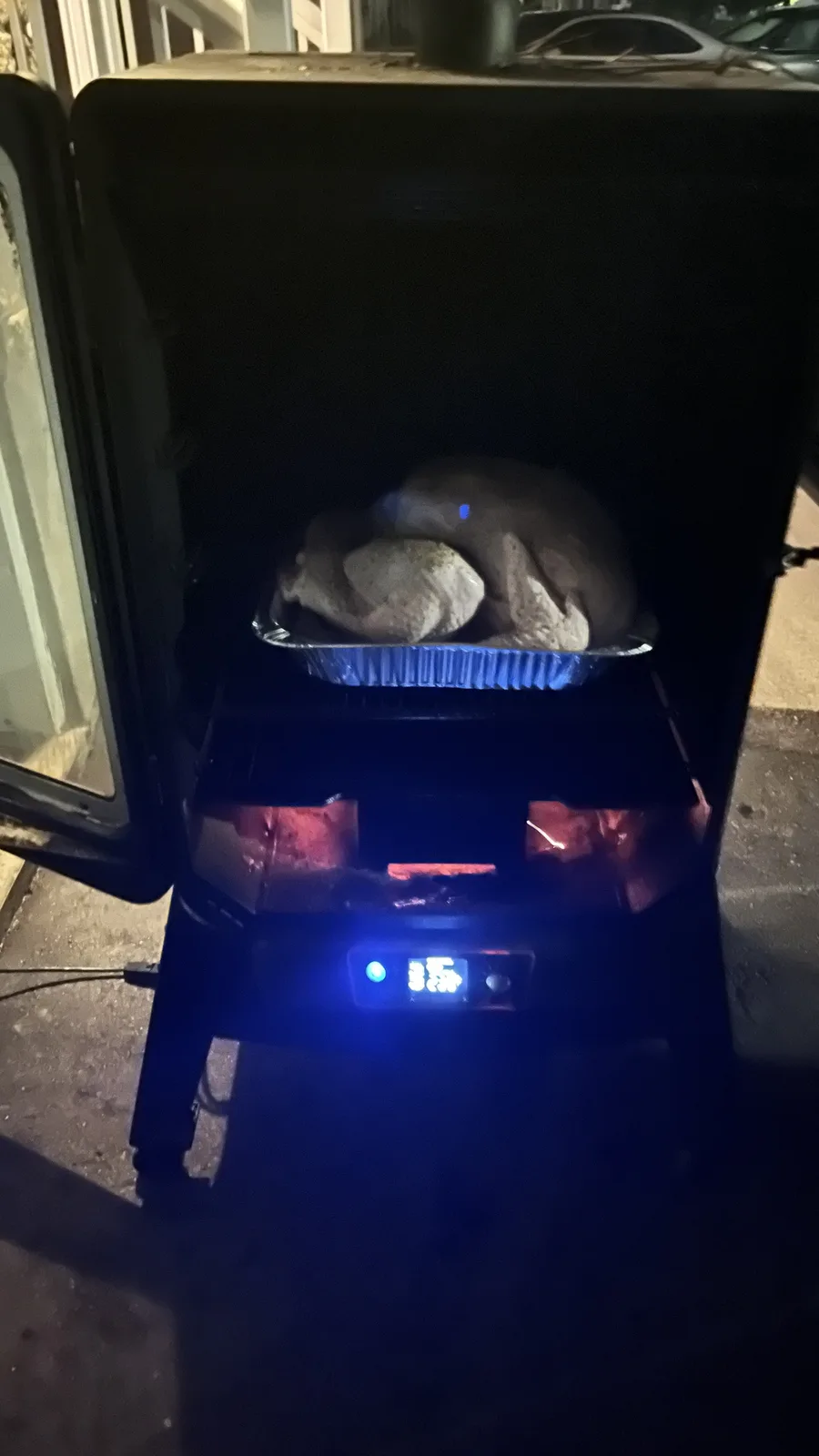 A whole raw turkey sits in a foil pan inside an open electric smoker at night, with glowing heating elements visible below…