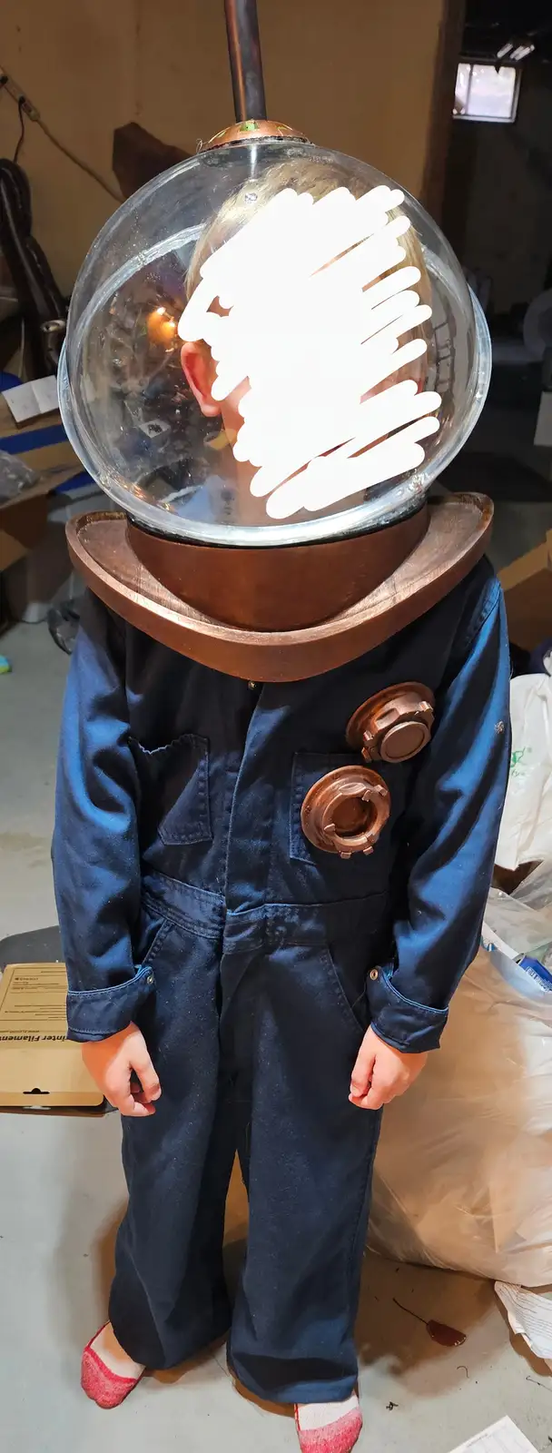A young child stands in a cluttered garage wearing a homemade deep-sea diving suit with a large glass helmet and copper…