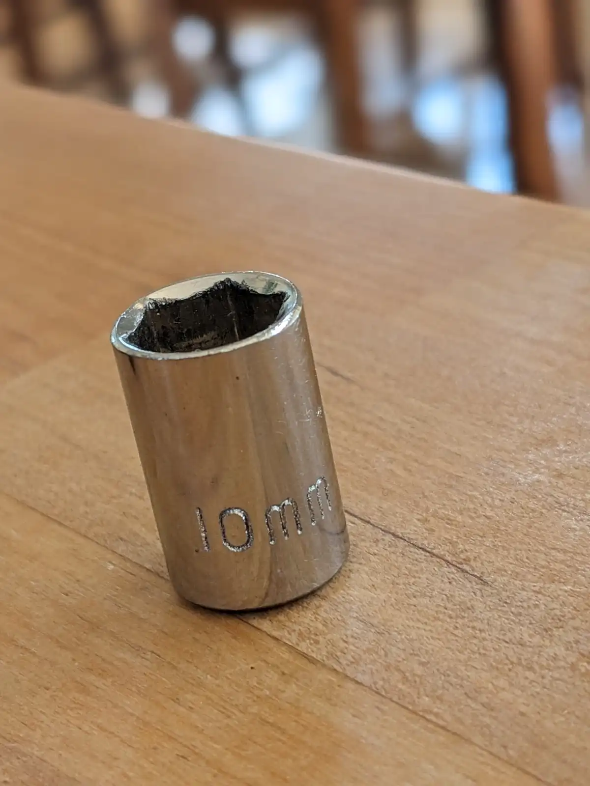 A close-up photo shows a worn, metallic 10mm socket wrench bit standing upright on a light brown wooden table. The socket's…