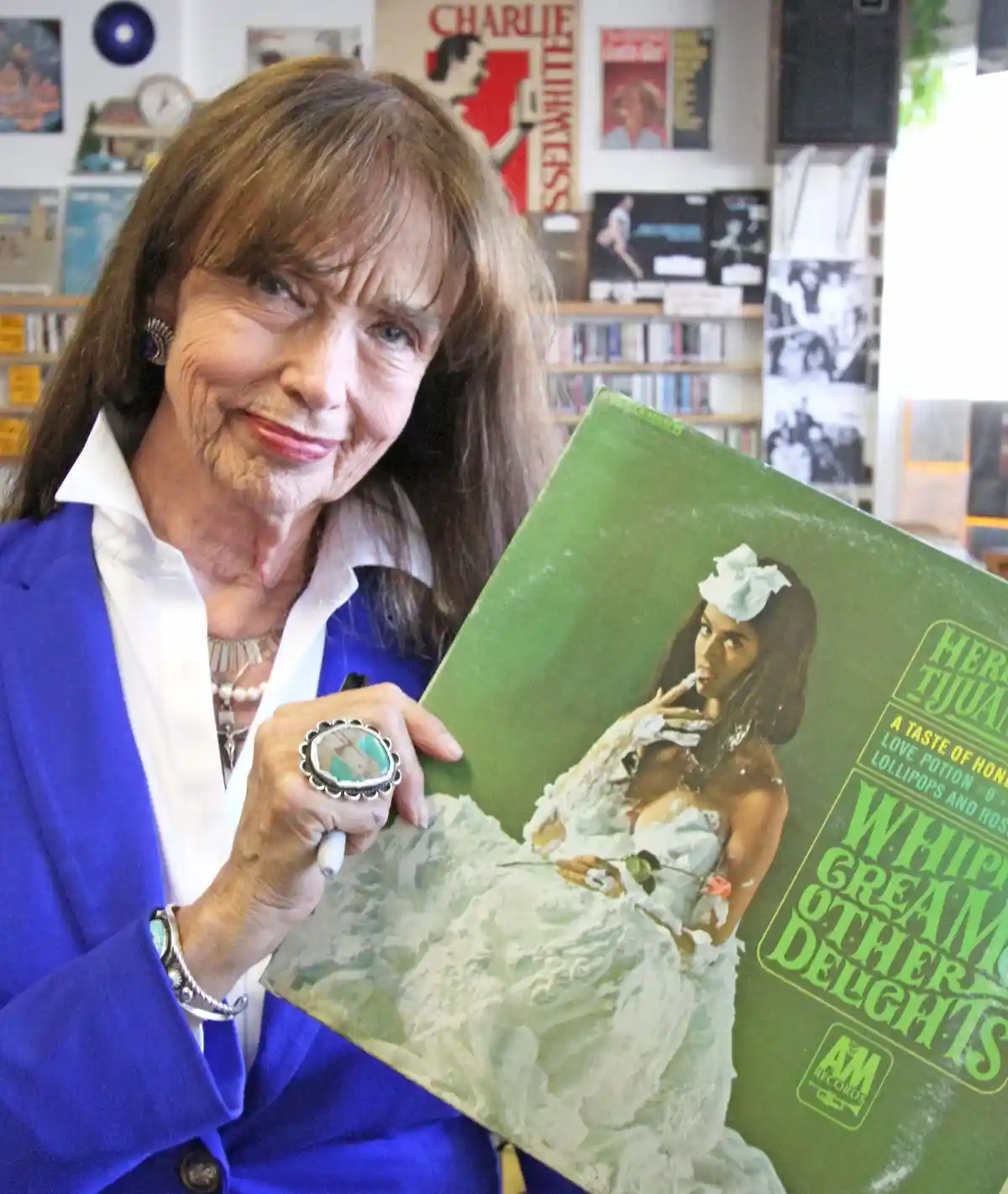 An older woman with brown hair and a blue blazer holds a green vinyl record album cover that has been covered in white…