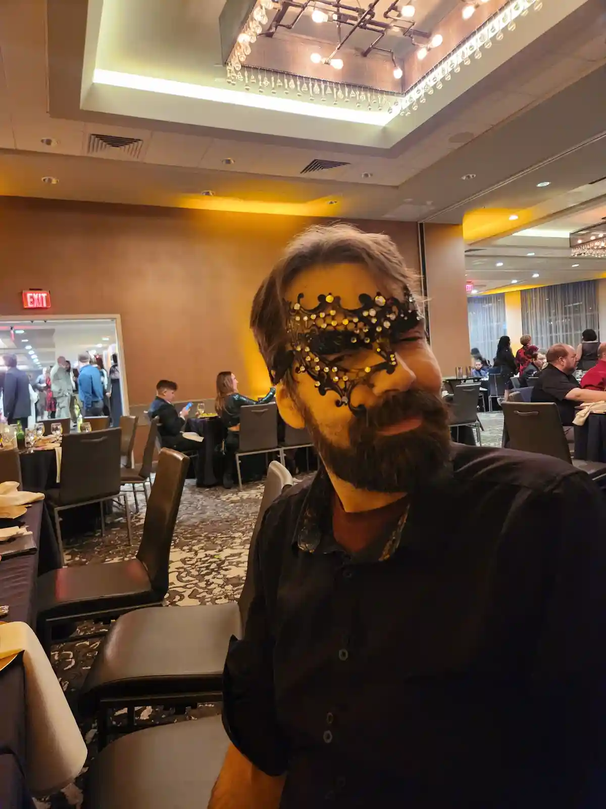 A man with a beard and a black ornate Venetian-style mask covering his eyes sits at a banquet hall table, smiling slightly…