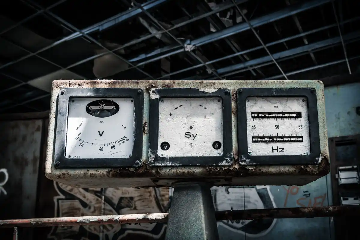 A close-up of a rusty, weathered industrial panel with three analog gauges: a voltmeter (V), a synchronization meter (Sy)…