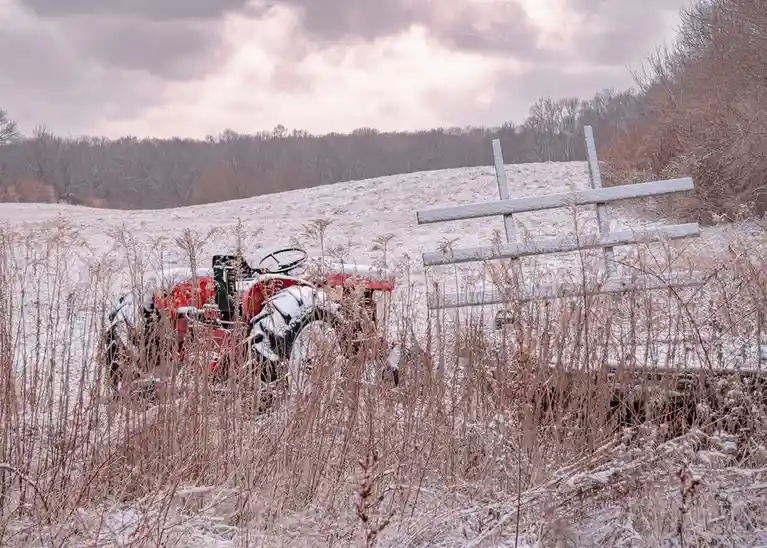 A red tractor sits abandoned in a snowy field, partially obscured by tall, dry grasses. A wooden fence lies toppled nearby…