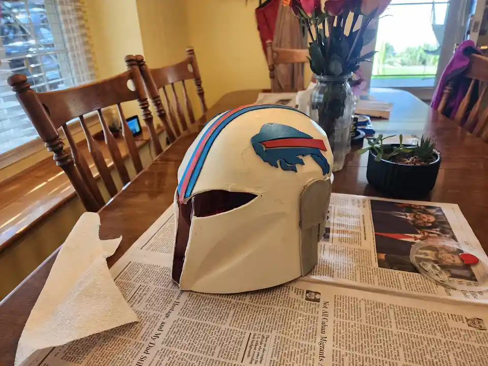 A white Mandalorian-style helmet with a red and blue stripe sits on a newspaper-covered dining table. The front of the…