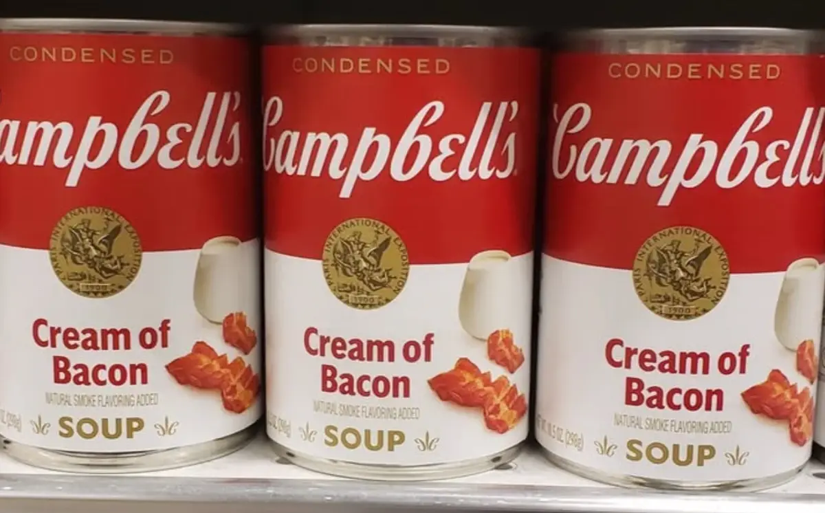 Three identical cans of Campbell's condensed soup are lined up on a shelf, each labeled 'Cream of Bacon' with 'Natural Smoke…