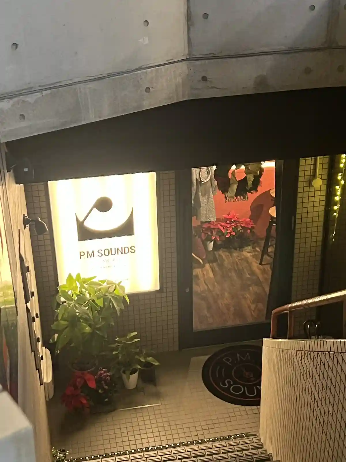 A photo of the entrance to a bar named 'P.M. SOUNDS' with a lit sign, potted plants, and a doormat featuring the same logo…