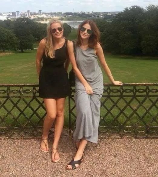 Two women stand side-by-side outdoors, leaning on a decorative metal fence. One wears a black dress and the other a grey…