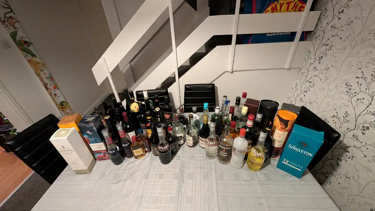 A table is covered with dozens of liquor bottles and boxes, including recognizable brands like Laphroaig, Talisker, Jameson…