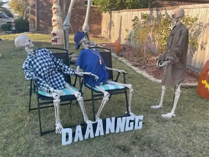 Three skeleton decorations are arranged in a backyard Halloween scene. Two skeletons sit in folding chairs, one wearing a…