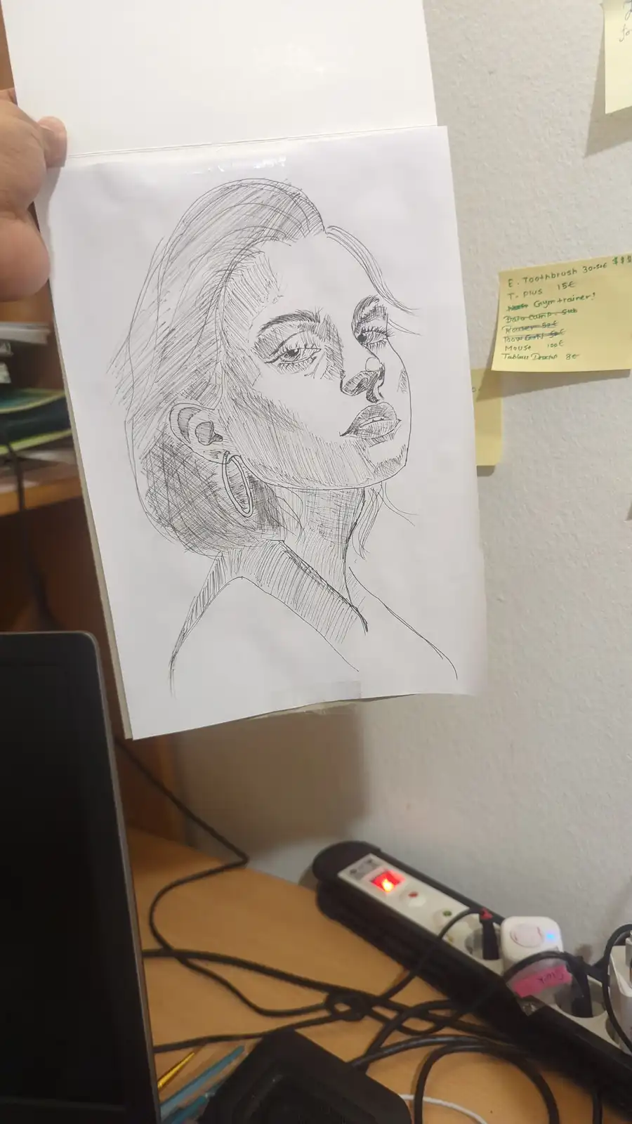 A hand holds up a detailed pencil sketch of a woman's face, showing her profile with defined features and hoop earrings. In…