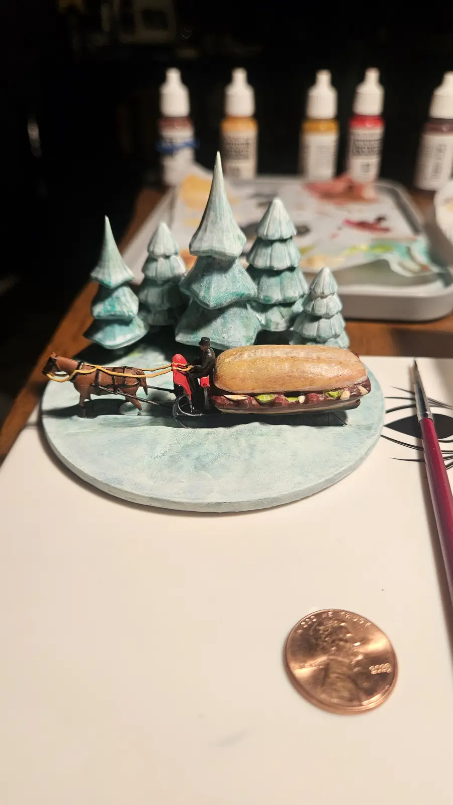 A miniature diorama shows a tiny horse-drawn sleigh pulled by a brown horse, with a miniature figure in a red coat driving…