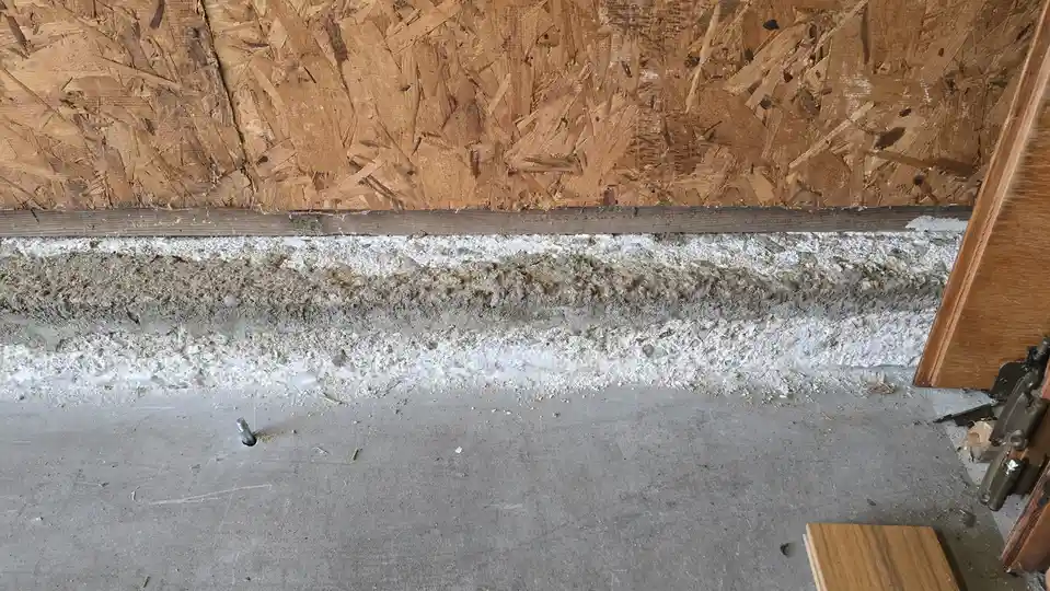A close-up photo shows a concrete floor meeting a wall with OSB paneling; a dirty, crumbling white and gray horizontal band…