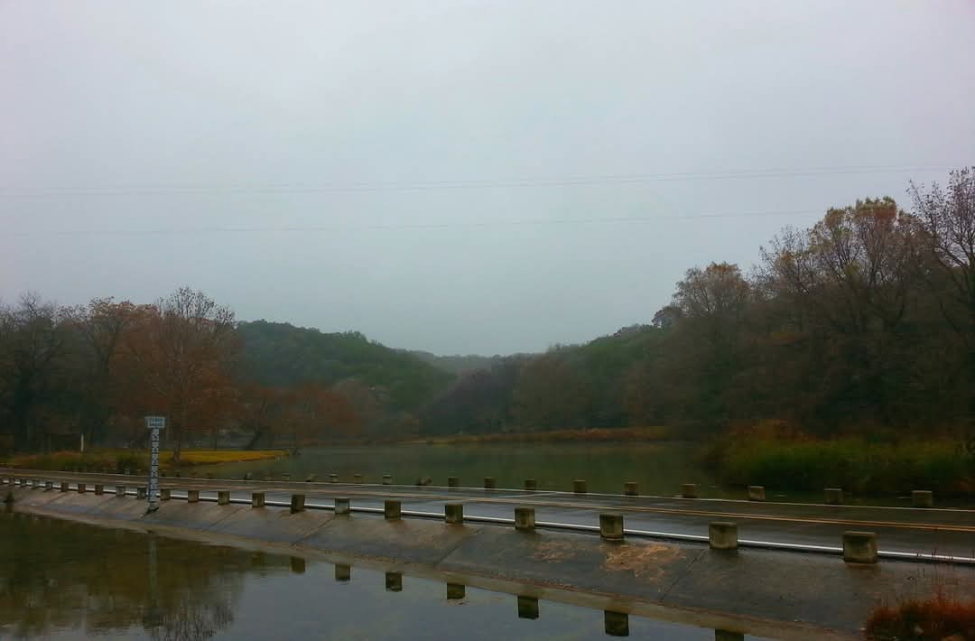 A gloomy, overcast landscape featuring a wet road with a guardrail running alongside a body of water, with trees and hills…