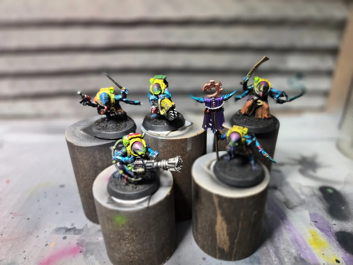 Five brightly painted fantasy miniatures, each standing on a wooden dowel, are arranged on a paint-splattered surface. The…