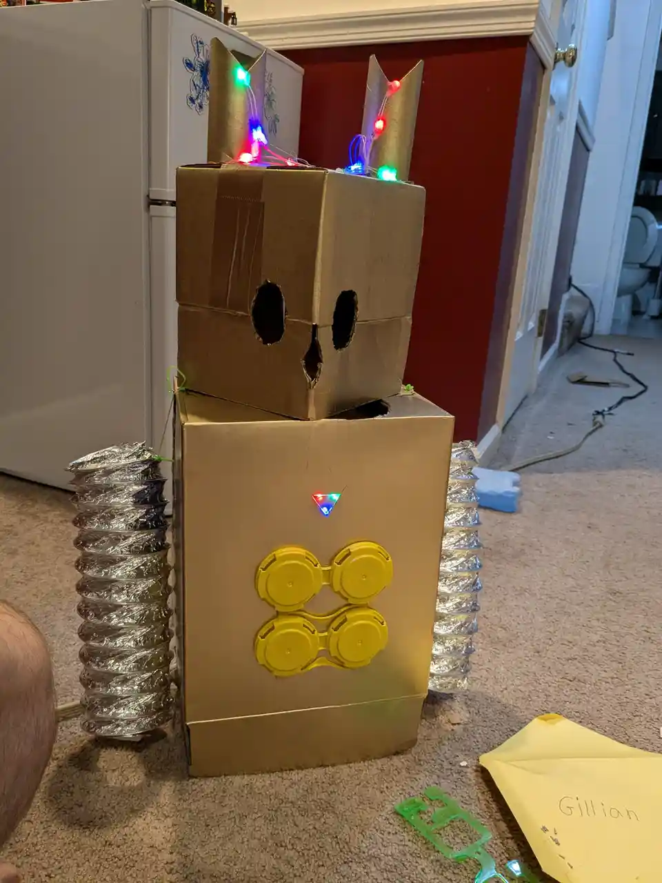 A homemade cardboard robot costume stands on a carpeted floor, constructed from two stacked boxes. The head box has two eye…