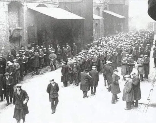 A black-and-white historical photograph shows a large crowd of men in early 20th-century attire, mostly wearing hats and…