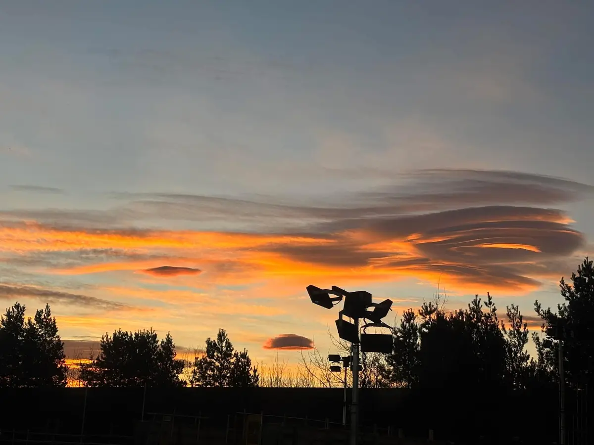 A photograph of a dramatic sunset sky with vibrant orange and pink clouds, featuring a large, wavy, lens-shaped cloud…