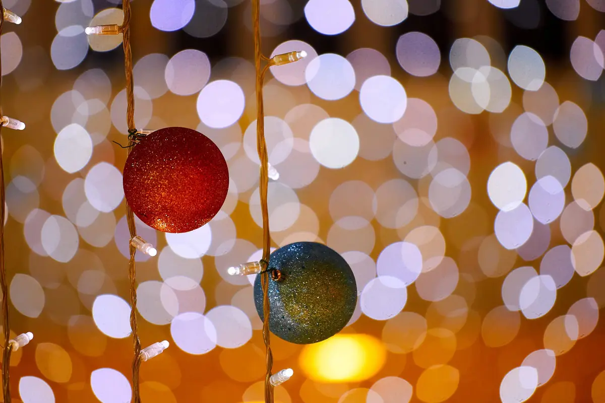 A close-up photo shows two glittery Christmas ornaments, one red and one blue-green, hanging on a string of warm white fairy…