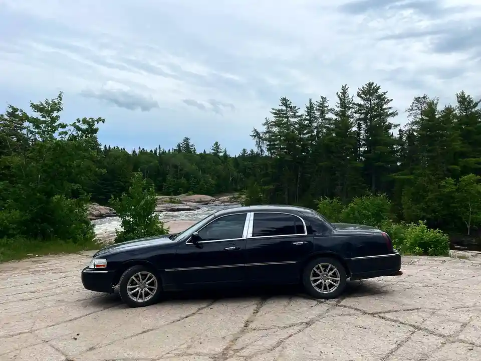 A black luxury sedan, resembling a limousine, is parked on a cracked concrete surface beside a rushing river with dense…