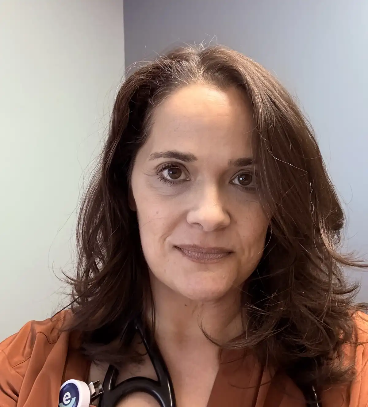 A woman with brown hair and a stethoscope around her neck, wearing an orange top, looks directly at the camera with a slight…