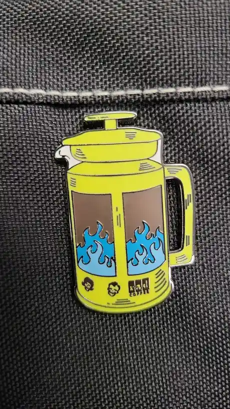 A yellow enamel pin shaped like a French press coffee maker, featuring cartoonish blue flames rising from brown coffee…