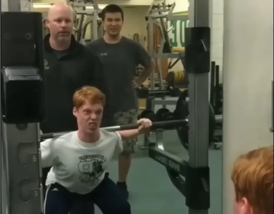 A young man with red hair is performing a squat with a barbell in a gym, his face contorted in intense effort. Two men stand…