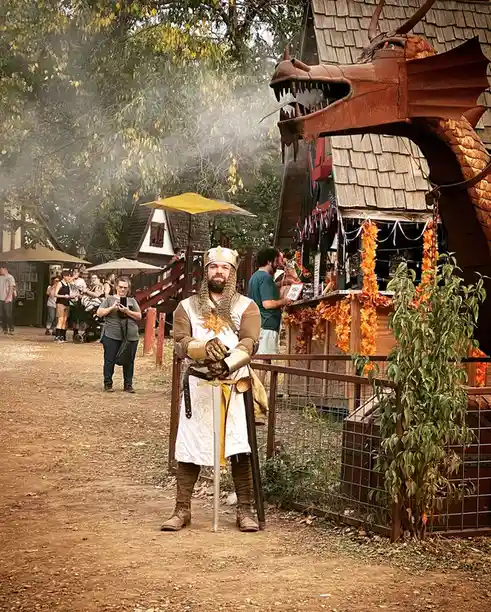 A man in a medieval knight costume, complete with chainmail, crown, and sword, stands confidently in front of a large…