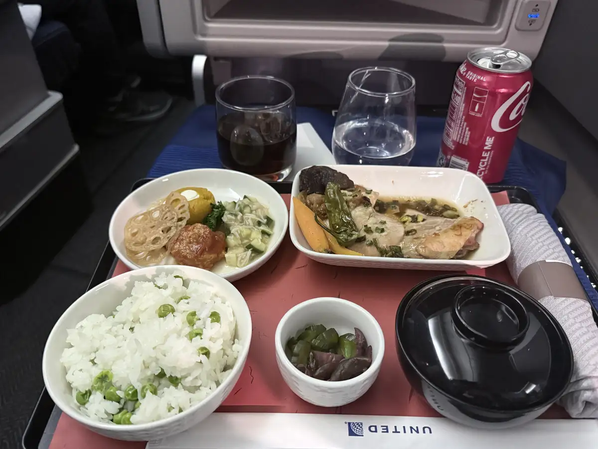 A close-up photo of a United Airlines in-flight meal tray featuring a bowl of rice with peas, a main dish with chicken and…