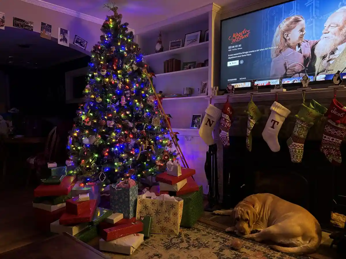 A cozy, dimly lit living room is decorated for Christmas with a brightly lit tree, stacks of wrapped gifts, and stockings…