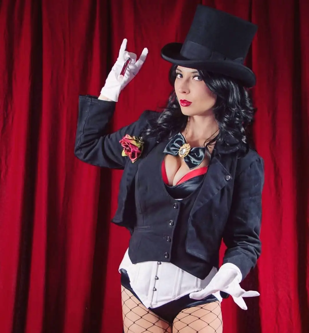 A woman dressed as a magician in a black top hat, black jacket, white gloves, and fishnet stockings stands before a red…
