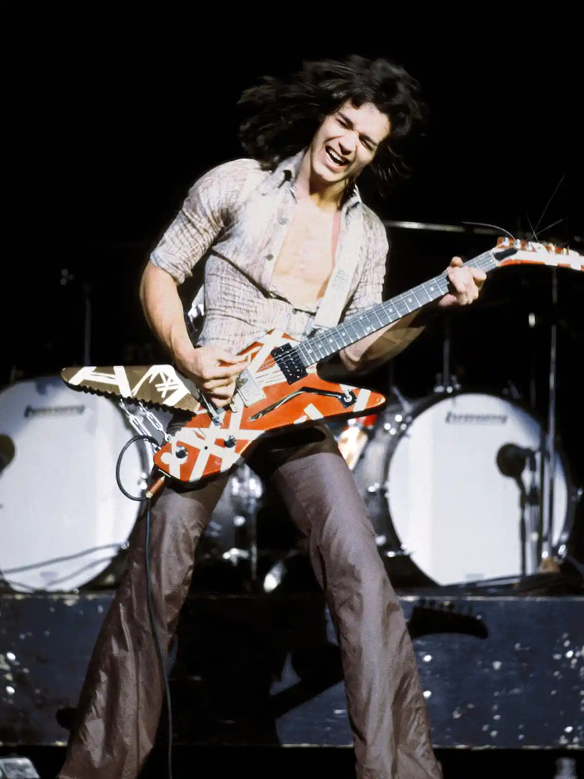 A man with long dark hair, wearing a light-colored shirt and dark pants, is energetically playing a distinctive orange and…