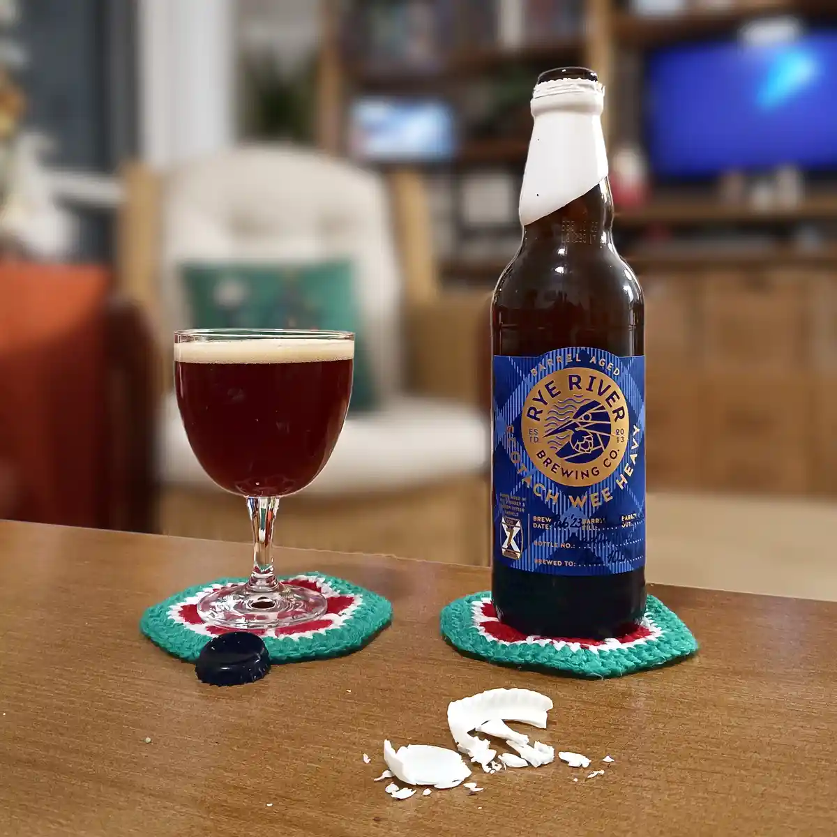 A dark beer bottle and glass sit on a wooden table, both resting on colorful crocheted coasters. The bottle, labeled 'Rye…