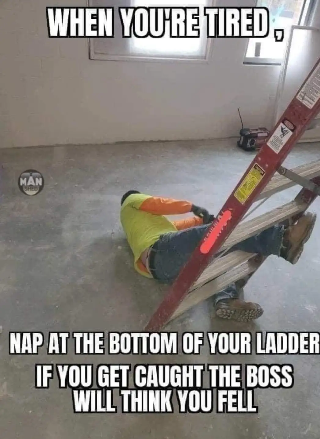 A person in a yellow and orange work shirt and jeans is lying on a concrete floor, using the base of a red ladder as a…