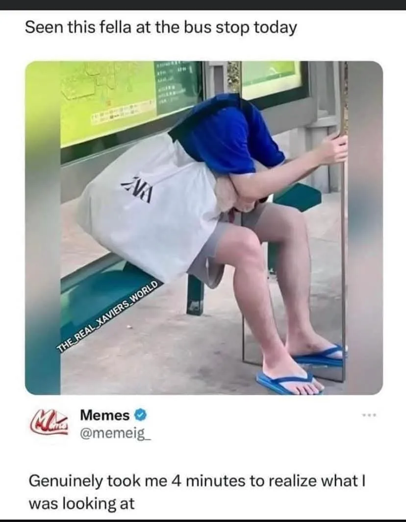 A person sits on a bus stop bench, leaning forward with their head down, wearing a blue shirt, grey shorts, and blue…