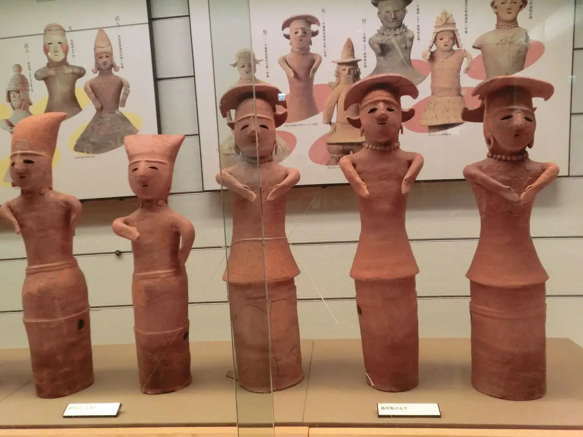 A row of ancient Japanese clay figurines, likely from the Yayoi or Kofun period, displayed in a museum case. They have…