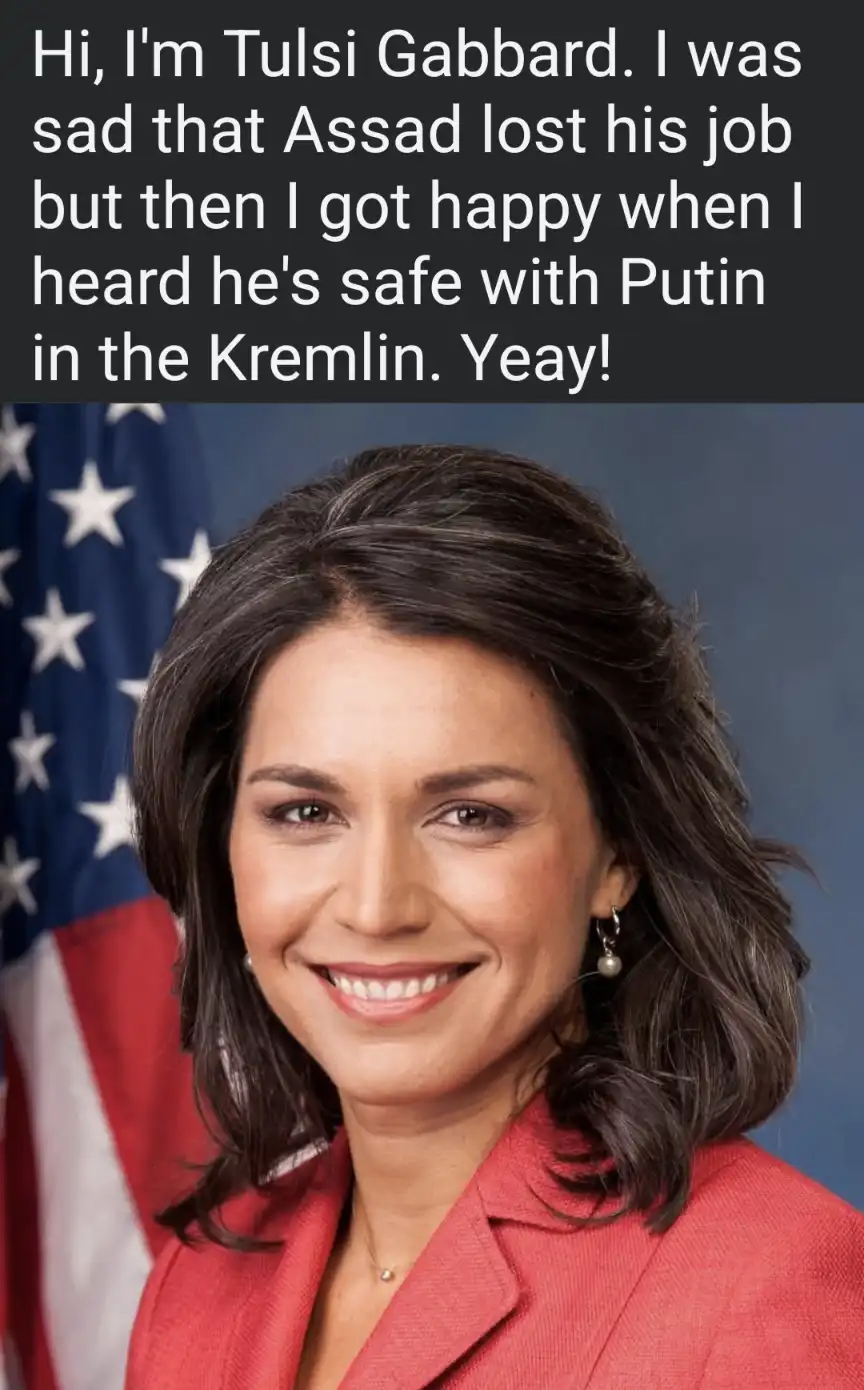 A formal portrait of Tulsi Gabbard smiling, wearing a red blazer with an American flag visible behind her left shoulder. The…