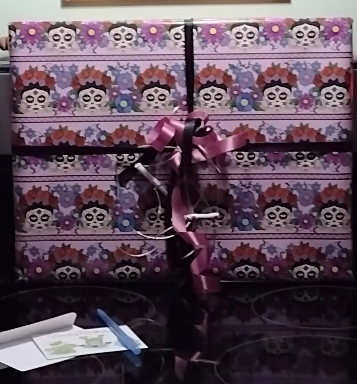 A gift wrapped in purple sugar skull patterned paper with a pink ribbon is sitting on a black stovetop. A white pen and a…
