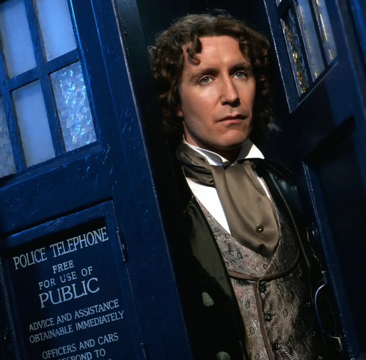 A man with curly brown hair and period clothing stands in the doorway of a blue British police box, which has a sign reading…