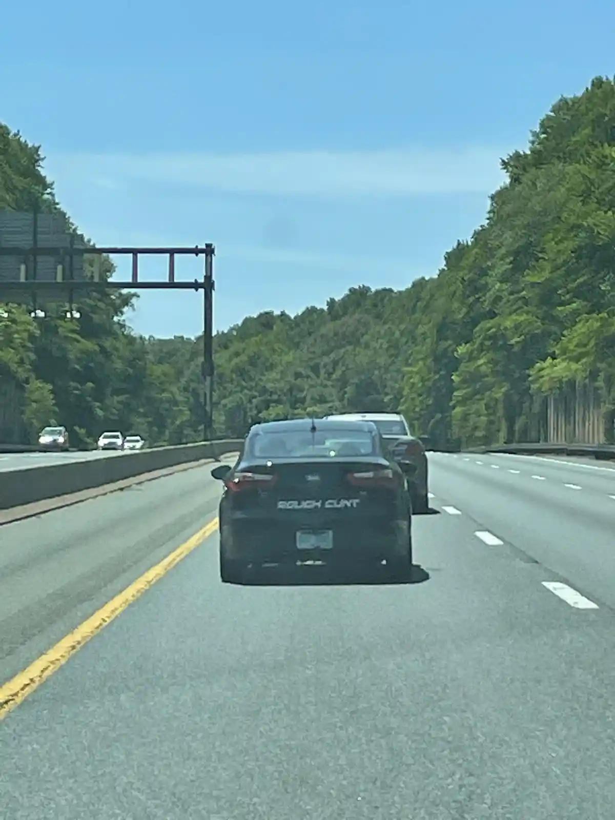 A black sedan is driving on a multi-lane highway with trees on both sides under a clear blue sky. The car's rear trunk has…