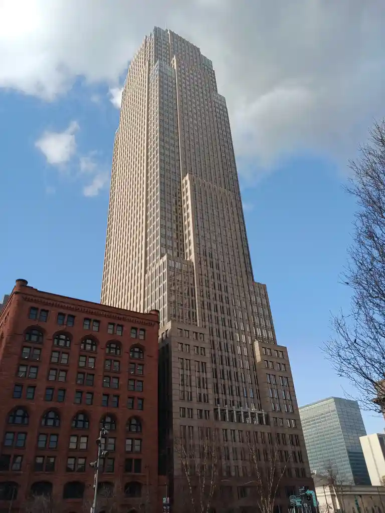 A low-angle photo shows a tall, modern skyscraper with a beige facade and many windows, towering over a smaller red-brick…