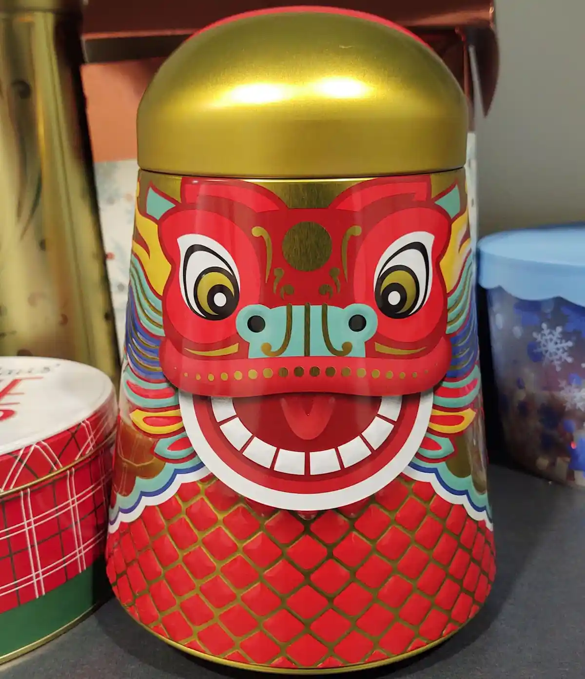 A close-up photo of a decorative red and gold cylindrical tin with a large, cartoonish lion dance mask design on its side…