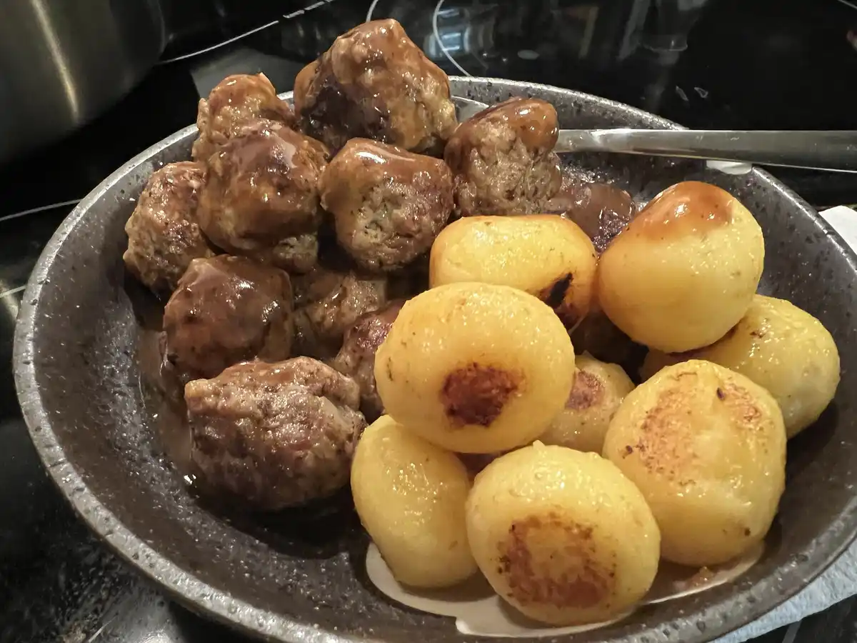 A close-up photo shows a dark speckled bowl filled with brown, sauce-covered meatballs and golden-brown roasted potatoes. A…