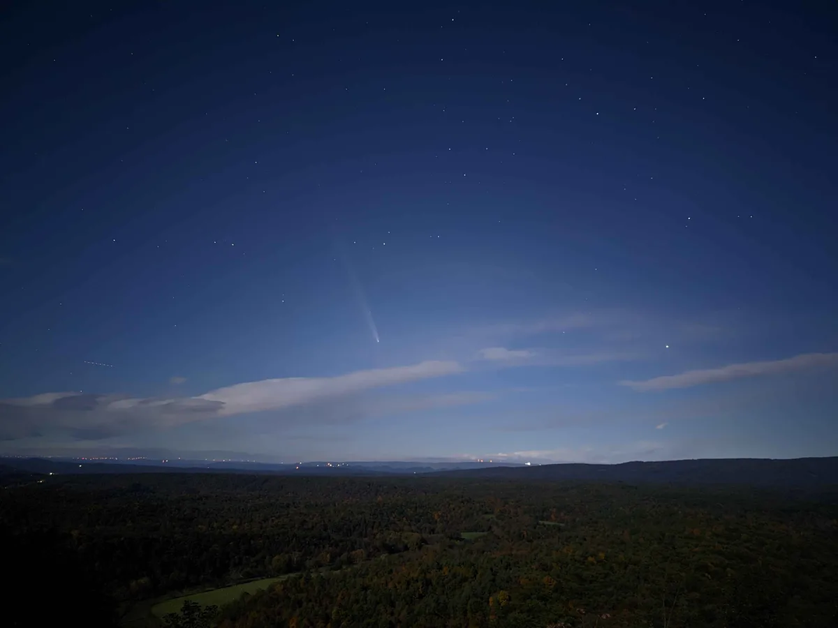 A night sky photograph shows a bright comet with a visible tail streaking across a starry sky above a forested mountain…