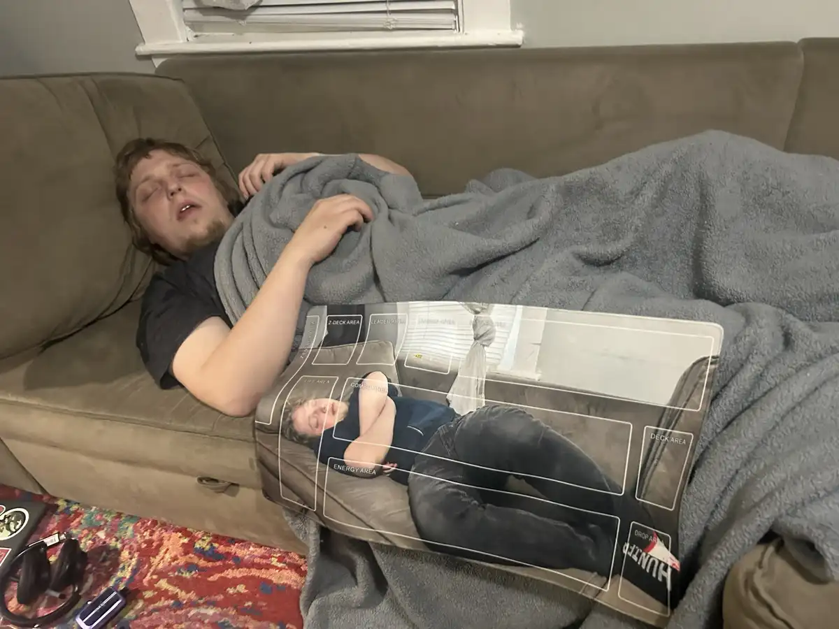 A person is sleeping on a couch under a gray blanket, with a transparent overlay showing a game board labeled with areas…