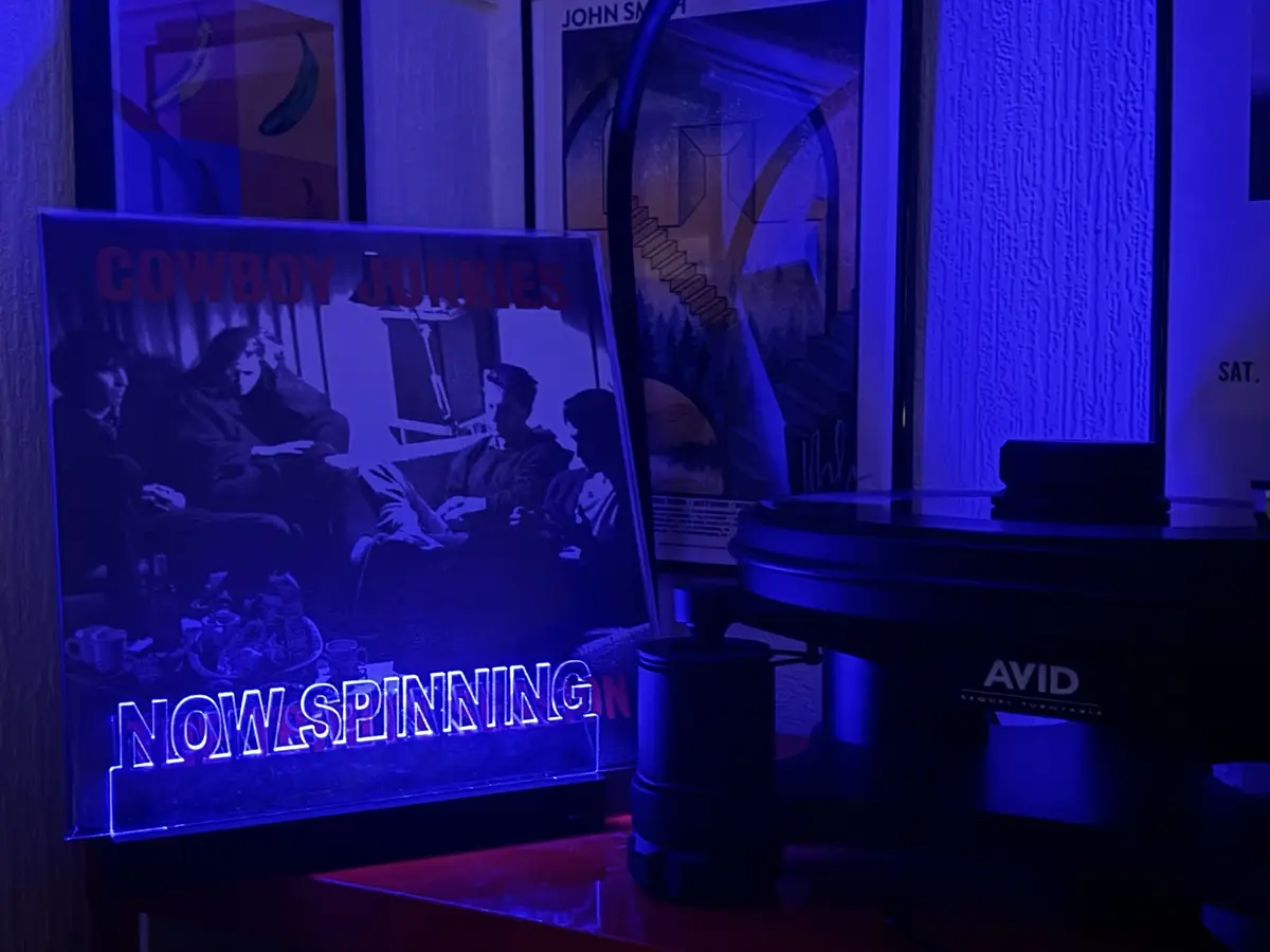 A dimly lit room bathed in blue light features a vinyl record player with an 'AVID' logo, next to a glowing neon sign…