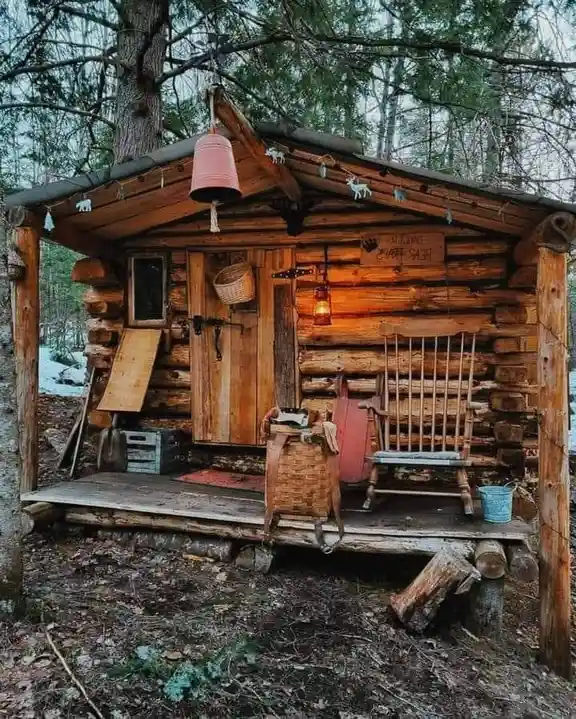 A rustic log cabin in a snowy forest with a porch featuring a rocking chair, a basket, and a lantern. A pink bell hangs from…