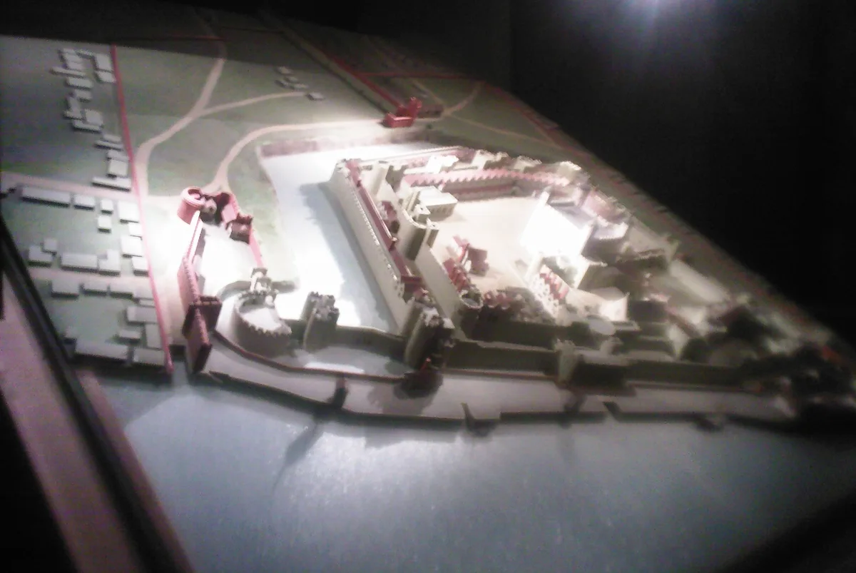 a blurry, low-light photograph of a physical model of a castle with moat and walls, displayed under a spotlight. The…