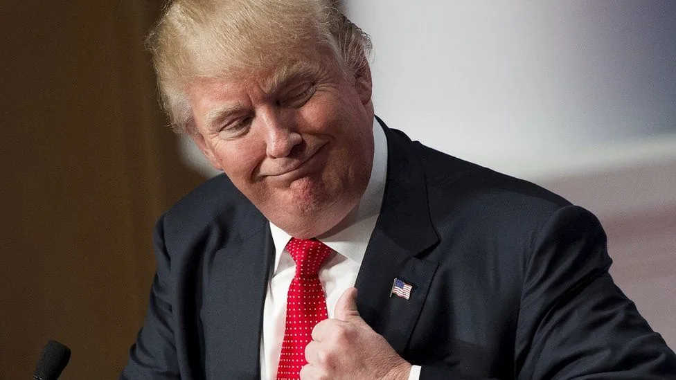 A man with blond hair, wearing a dark suit, white shirt, and red polka-dot tie, gives a thumbs-up while smiling and looking…